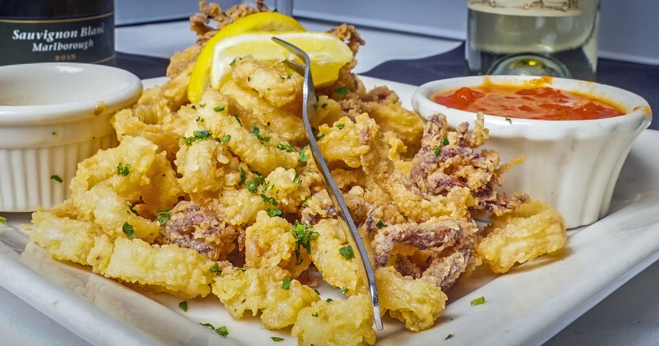 A plate of fried calamari with lemon and two dipping sauces beside a bottle of white wine