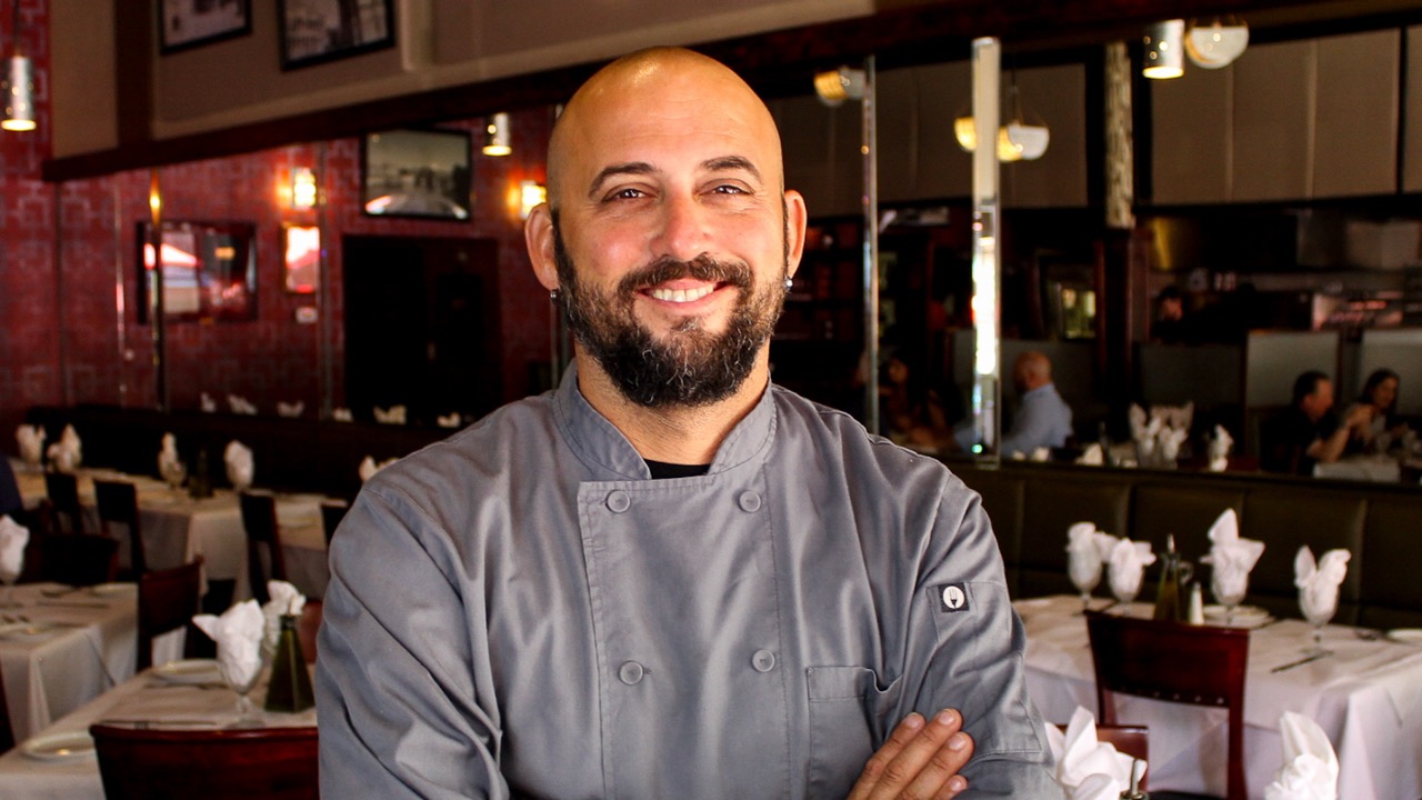 A smiling chef in a gray chef’s coat stands with arms crossed in the Gratzzi dining room