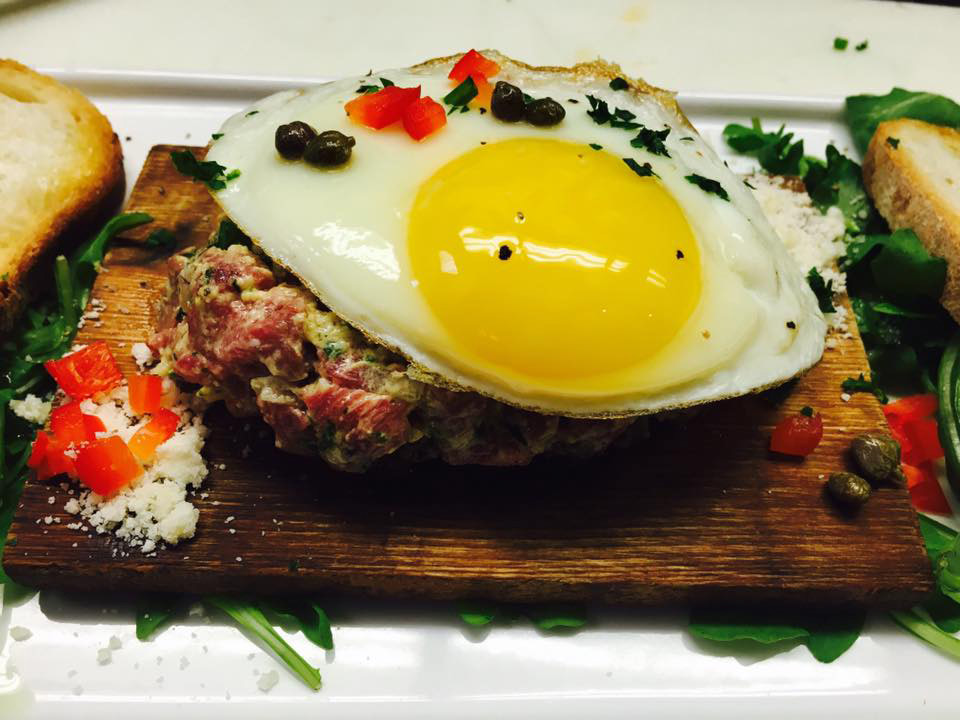Steak tartare on a wooden plank under a fried egg with capers and toast points